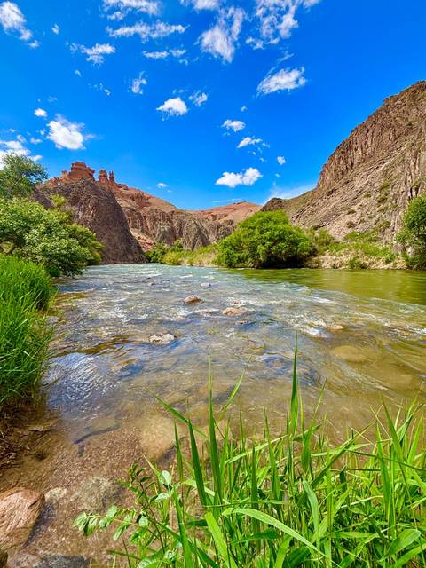       Clear mountain river rushing between green banks and red sandstone canyon walls beneath bright blue sky.
  