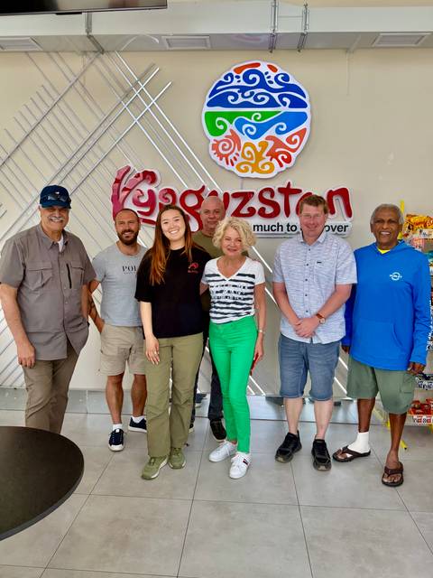       Group of smiling travellers posing in front of a colourful Kyrgyzstan promotional sign indoors.
  