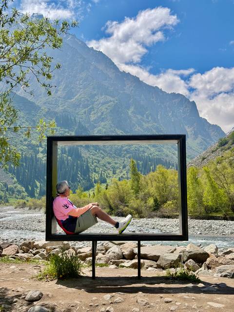       A man relaxes inside a large rectangular frame beside a clear river with lush green mountains towering behind.
  