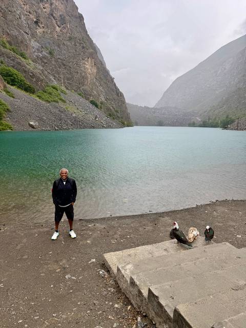       A lone traveler stands beside a turquoise alpine lake surrounded by rocky slopes under misty skies.
  