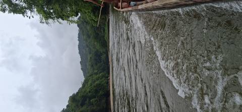      View from a wooden canoe speeding along a wide jungle river under overcast skies.
  