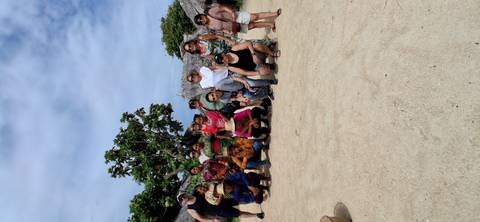       Tourists and local islanders posing together on a sandy beach beneath a bright sky.
  