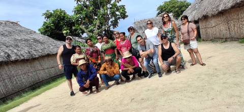       Tilted snapshot of travelers and locals gathered outside thatched huts on sandy ground.
  