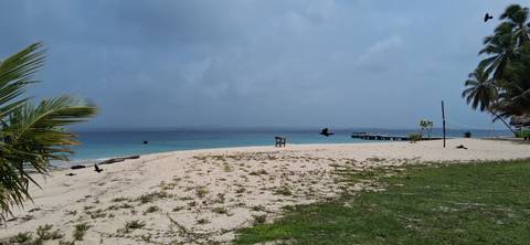       Quiet sandy island beach edged by calm turquoise water, a wooden pier and a few birds in flight under hazy skies.
  