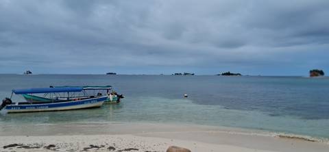       Small motorboat anchored in shallow clear water off a sandy shore with low islands on the horizon under grey clouds.
  