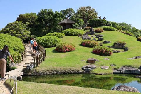      Manicured Japanese garden with stone paths, blooming shrubs and visitors crossing a bridge
  
