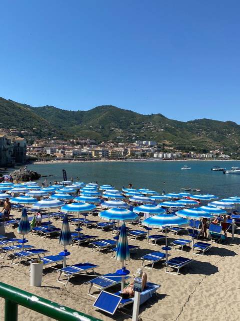       Mediterranean beach scene with rows of blue-striped umbrellas, boats offshore, and green hills behind a coastal town.
  