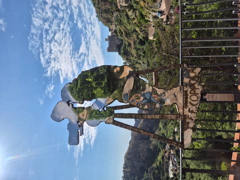       Reflective metal silhouette sculpture of a film director with Savoca hillside and castle in the background.
  