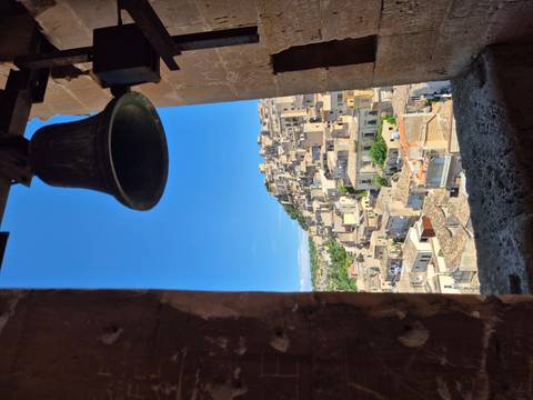       Picturesque hill town of Modica viewed through a bell-tower window with stone bell in silhouette.
  
