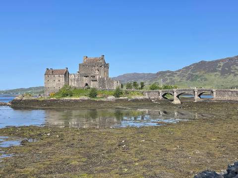       Stone Eilean Donan Castle sits on a tidal island reflected in calm waters with a small bridge.
  