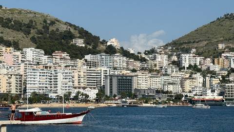       Dense seaside city of white apartment blocks backed by steep green hills and blue bay with a small boat.
  