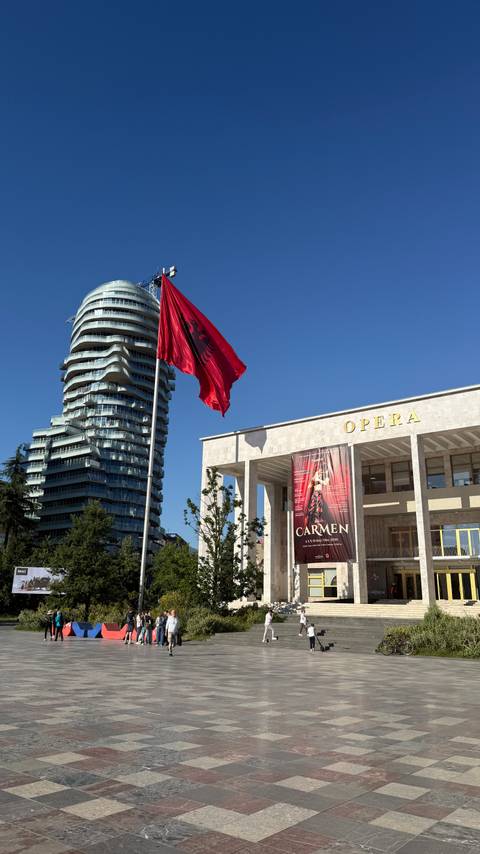       Modern opera house facade with a tall curving skyscraper and Albanian flags under clear sky.
  