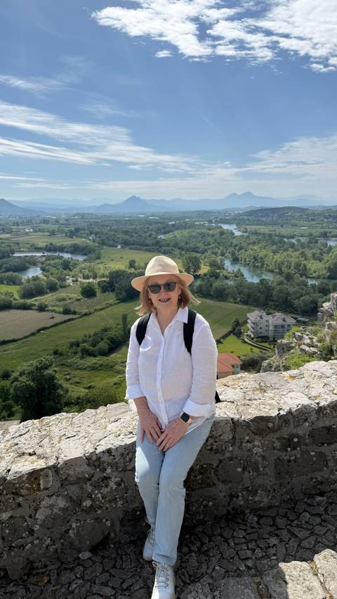       Smiling woman in hat and sunglasses poses with a lush river valley and farmland stretching behind her.
  