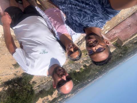       Three friends pose for a bright daytime selfie on a rocky hillside with distant plains behind.
  
