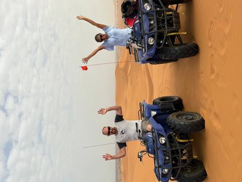       Two travelers on blue quad bikes raising their arms while parked on orange desert dunes under a lightly clouded sky.
  