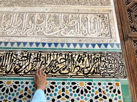      Close-up of ornate Arabic calligraphy carved in stone and colorful mosaic tiles with a hand touching the wall.
  