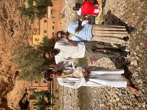       Three travelers posing on a rocky riverbed with tall ochre canyon walls and traditional buildings behind.
  