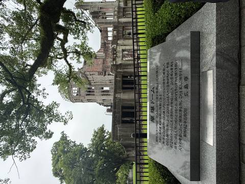       The ruined Atomic Bomb Dome framed by green trees behind a memorial stone in Hiroshima.
  