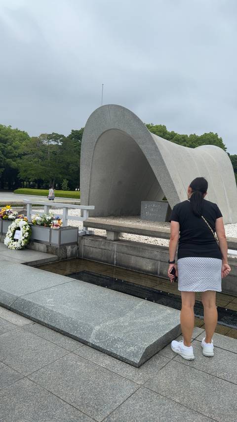       Visitor looking at the Cenotaph in Hiroshima Peace Park with floral wreaths nearby.
  