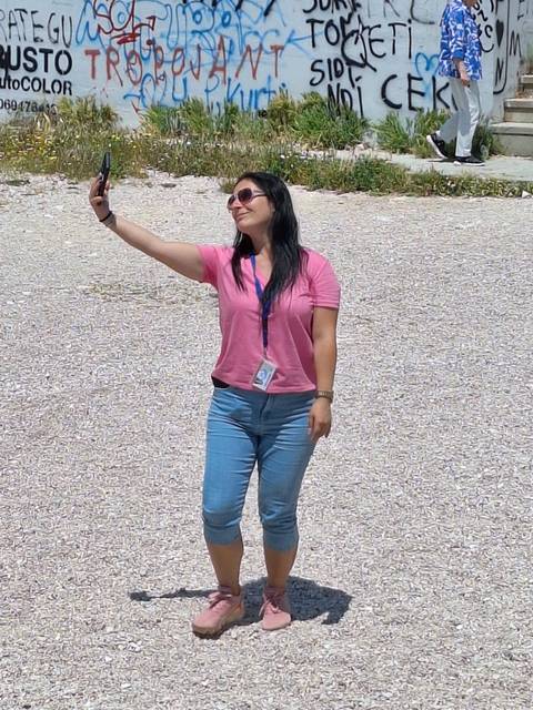       A woman in pink top takes a selfie against a gravel surface under bright sun.
  