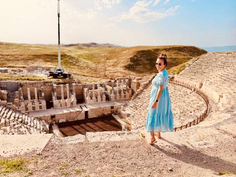       Traveller in a flowing blue dress stands above a sprawling ancient stone amphitheatre.
  