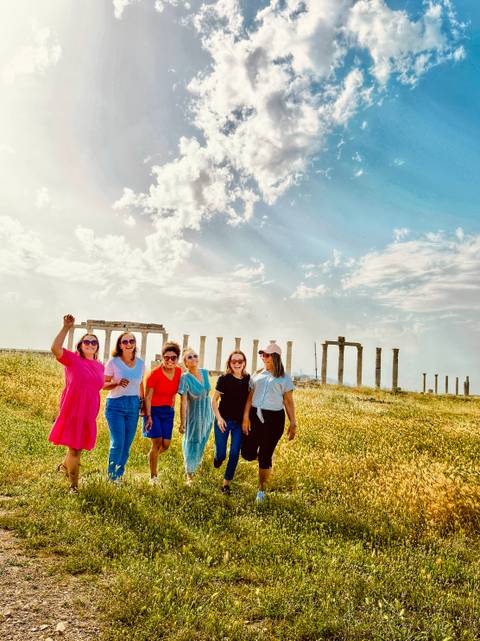       Group of friends pose cheerfully in a grassy archaeological field dotted with standing columns.
  