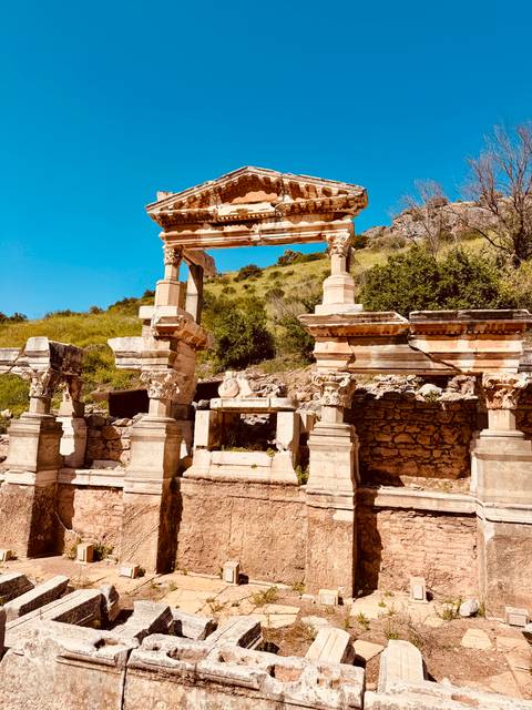       Sun-bleached ruin with decorative columns set against a green hillside and vivid blue sky.
  