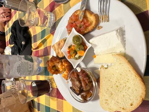       Plate of assorted Sicilian appetizers including bread, bruschetta, olives, cheese and fried rice ball on a colourful checked tablecloth at a restaurant.
  