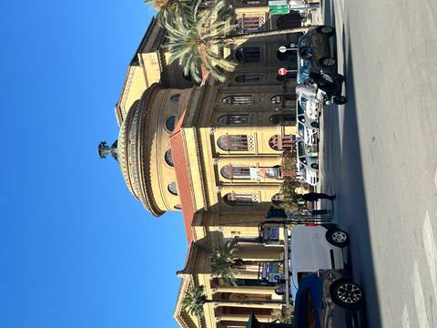       Teatro Massimo opera house in Palermo under a bright blue sky with traffic and pedestrians in front.
  