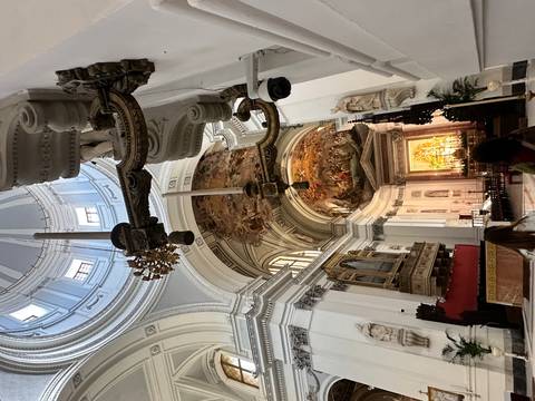       Baroque church interior with ornate ceiling frescoes, organ and visitors admiring the architecture.
  