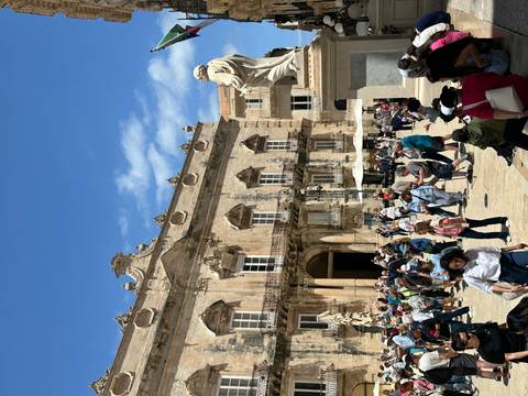       Busy piazza with baroque palace façade and marble statue surrounded by crowds of tourists under a sunny sky.
  