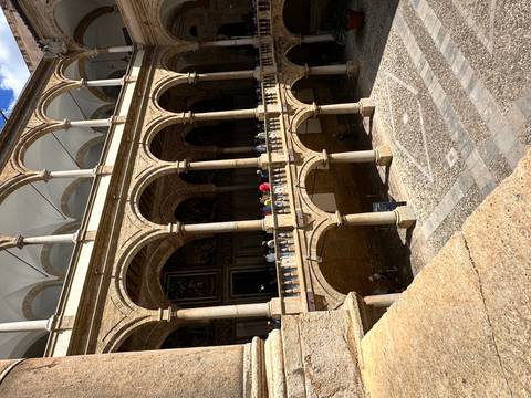       Stone cloister or palace courtyard with multiple arched levels and visitors walking along the balcony.
  