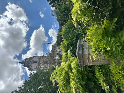       Baroque bell tower rising above lush green foliage and bright clouds in a hillside garden.
  
