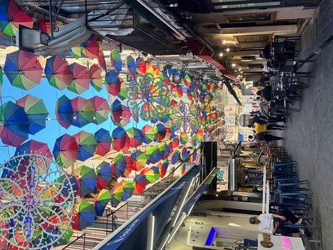       Narrow city street covered with a colourful canopy of hanging umbrellas, lined with cafés and shoppers.
  