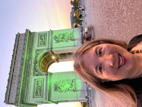       Woman taking an evening selfie with the illuminated Arc de Triomphe glowing green behind her.
  