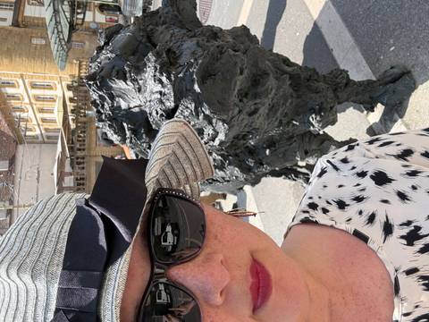       Close selfie of a woman in hat and sunglasses beside a dark metal animal sculpture on a sunny city street with historic yellow-stone buildings.
  