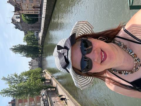       Woman taking a selfie on a bridge over a calm canal lined with leafy trees and old European buildings.
  