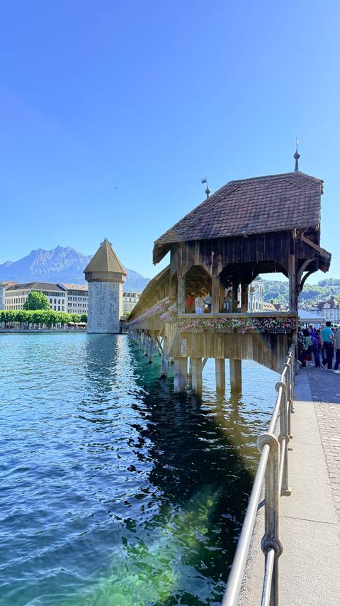       Iconic wooden Chapel Bridge with flower boxes crossing the turquoise river, medieval tower and alpine peaks behind.
  