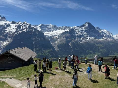       Crowds of tourists on an alpine viewpoint meadow overlooking dramatic snow-covered mountain peaks.
  