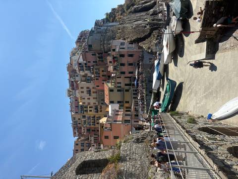       Colorful cliff-side village with small harbor, boats pulled onto a stone ramp and tourists walking along the waterfront under a bright blue sky.
  