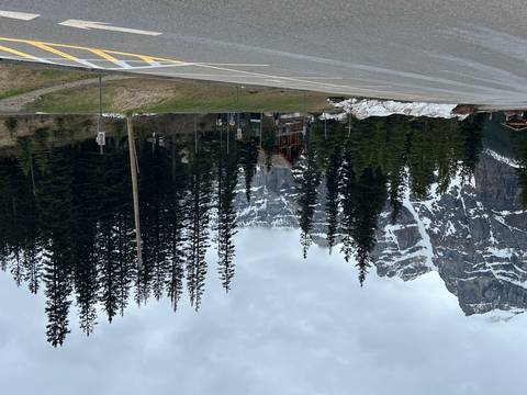       Mountain lodge nestled among tall pines with towering snow-streaked cliffs behind, viewed from a roadside parking area.
  