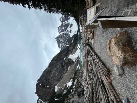       Shoreline of Moraine Lake with driftwood logs, turquoise water and dramatic snow-clad peaks under a gray sky.
  