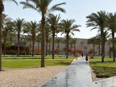       Landscaped gardens with palm trees outside a modern geometric-patterned museum in Giza.
  