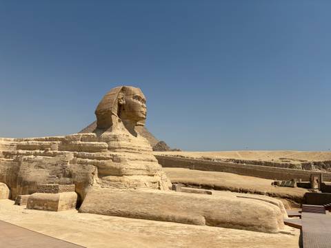       Side profile of the Sphinx with desert plateau and pyramid backdrop under clear blue sky.
  