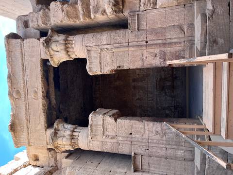       Entrance to a dimly lit ancient chamber flanked by carved columns with wooden steps leading up.
  