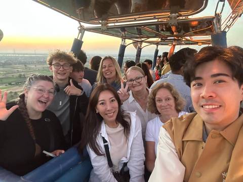      Happy travellers taking a selfie inside a hot-air balloon basket at dawn over farmland.
  