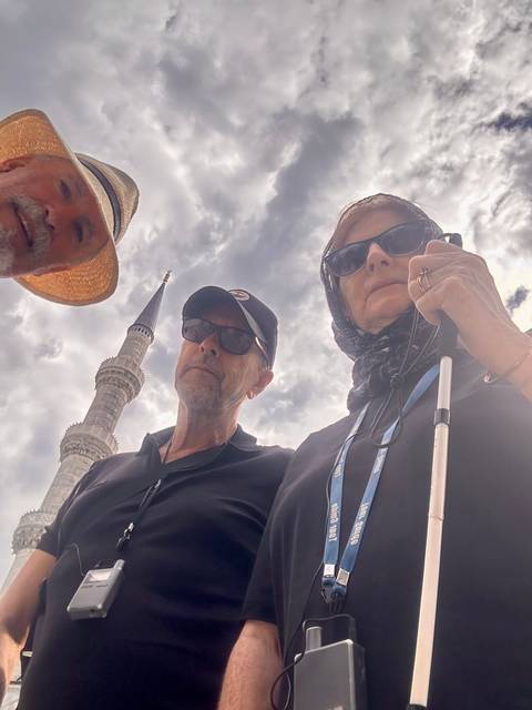       Three travellers posing beneath the soaring minaret of a mosque under dramatic clouds.
  