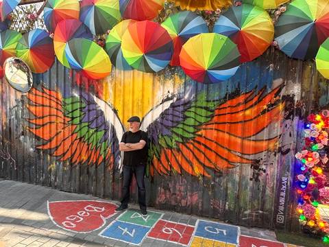       Man standing with arms crossed in front of a vibrant wing mural beneath hanging umbrellas.
  