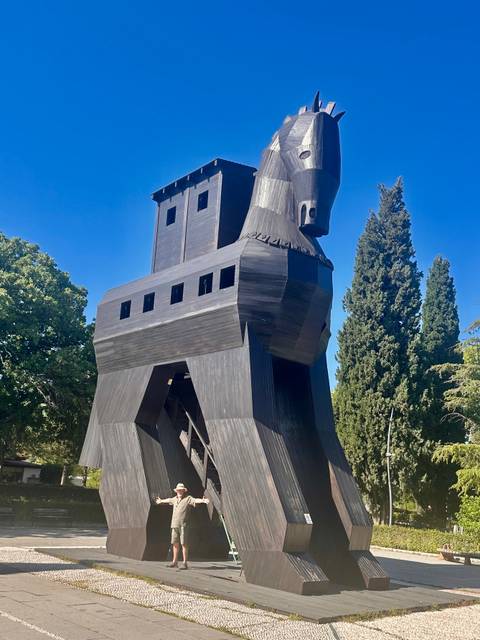       Large wooden replica of the Trojan Horse surrounded by tall evergreens under clear blue sky.
  