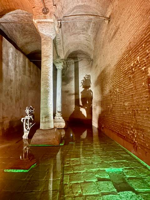       Moody interior of an ancient cistern showing columns and brick walls lit to reveal a Medusa head shadow.
  